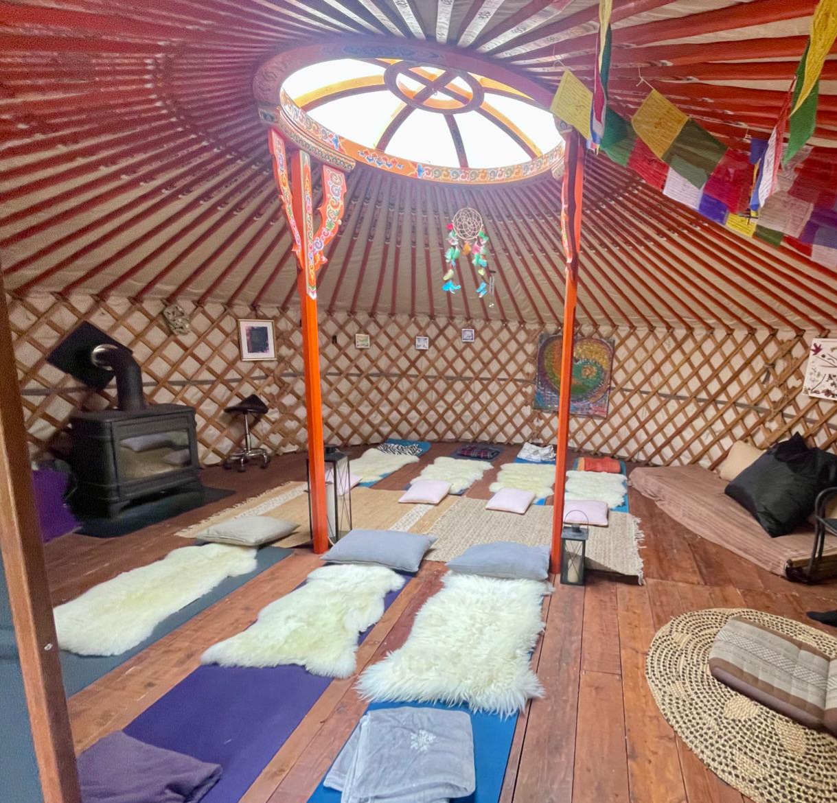 Intérieur chaleureux de la yourte : tapis de yoga, peaux de mouton, poêle à bois, drapeaux de prières et coupole lumineuse